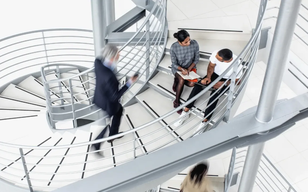 Large modern spiral staircase with a man walking up quickly, while two other people sit on the steps having a meeting.