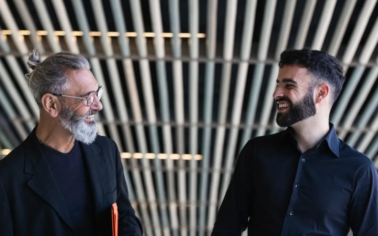 Two men facing each other, laughing, with a ceiling of elongated panels in the background.