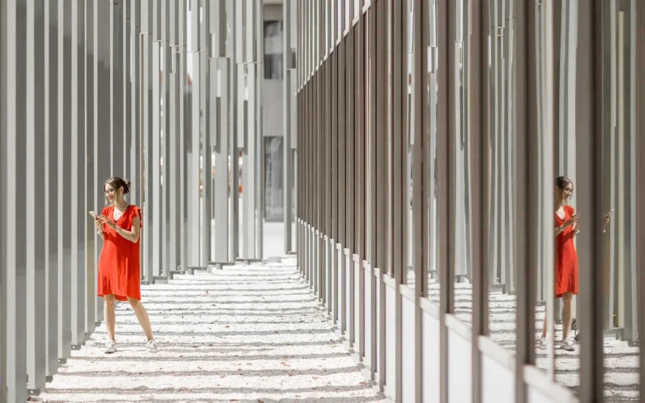 Woman in an orange-red dress holding a phone, standing in a passageway with architectural structures on both sides.