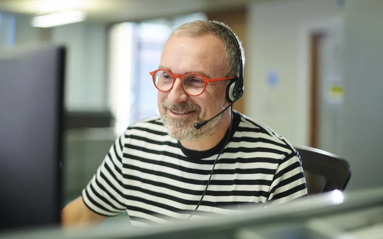 Man with red glasses and striped shirt sitting in front of a monitor, wearing a headset and engaged in conversation