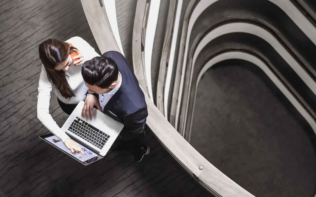 Bird's-eye view of a woman and man standing in a modern stairwell, looking at an open laptop screen together; a spiral staircase with a wooden railing is visible.