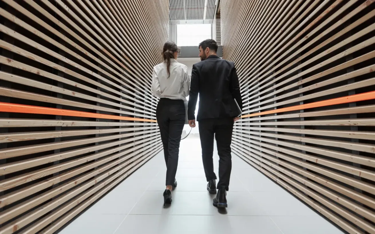 Woman and man walking down a hallway with wooden panels on the sides, each featuring an orange panel in the center