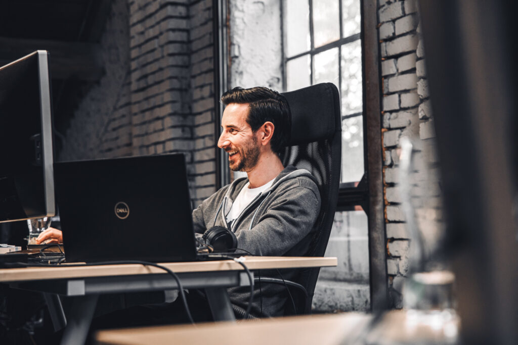 Ein Mann sitzt an einem Schreibtisch in einem Büro und lächelt, während er auf einen Computermonitor schaut. Er steht vor einer Backsteinmauer mit einem Fenster.