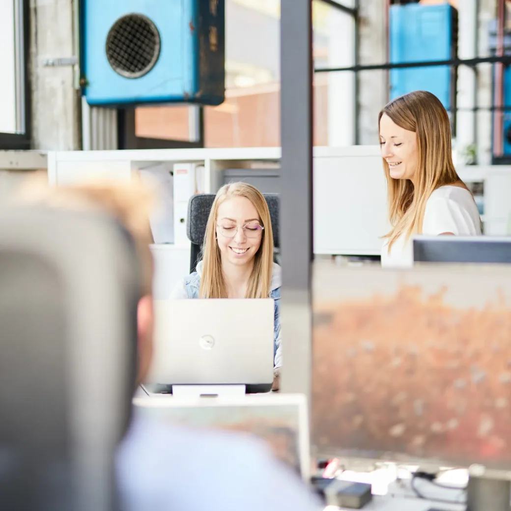 Zwei Frauen arbeiten gemeinsam an einem Schreibtisch in einem hell erleuchteten Büro. Eine sitzt mit einem Laptop, die andere steht und lächelt. Im Vordergrund ist eine verschwommene Person zu sehen.