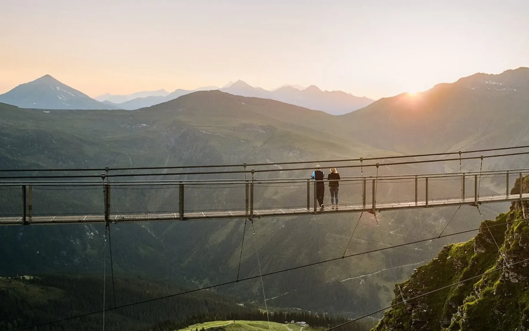 Bild von einer Hängebrücken in den Bergen von Skigastein, auf der zwei Personen stehen und in den Sonnenaufgang schauen.