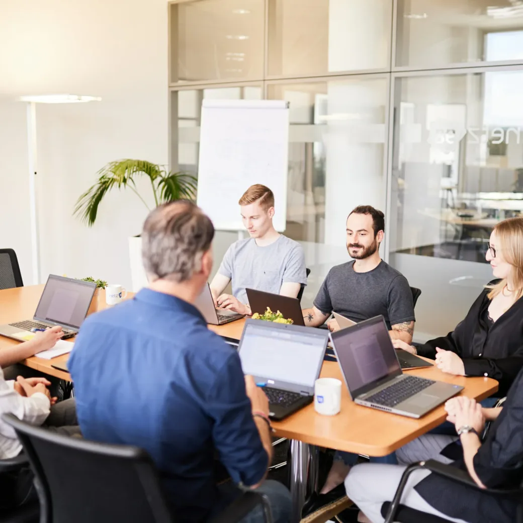 Eine Gruppe von sieben Personen sitzt mit Laptops und Dokumenten um einen Konferenztisch und nimmt an einer Besprechung in einem modernen Büroraum teil.