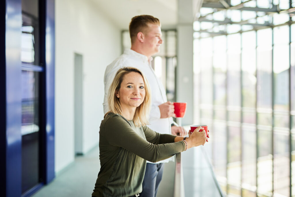 Ein Mann und eine Frau, die jeweils mit einer Tasse am Balkongelände ihrer Firma stehen.