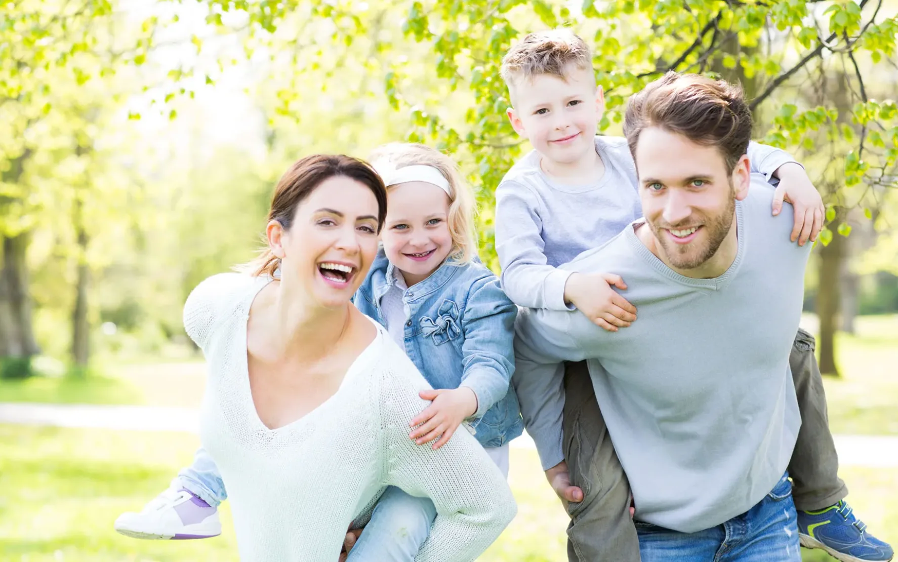 Bild von einer vierköpfigen Familie im Wald: Mutter und Vater tragen die Kinder huckepack und alle lachen in die Kamera.