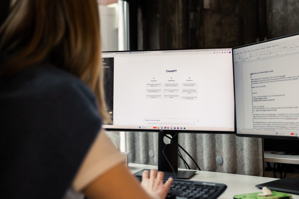 Eine Person sitzt an einem Schreibtisch mit zwei Monitoren und arbeitet an einem Computer. Der eine Bildschirm zeigt die Benutzeroberfläche von ChatGPT.
