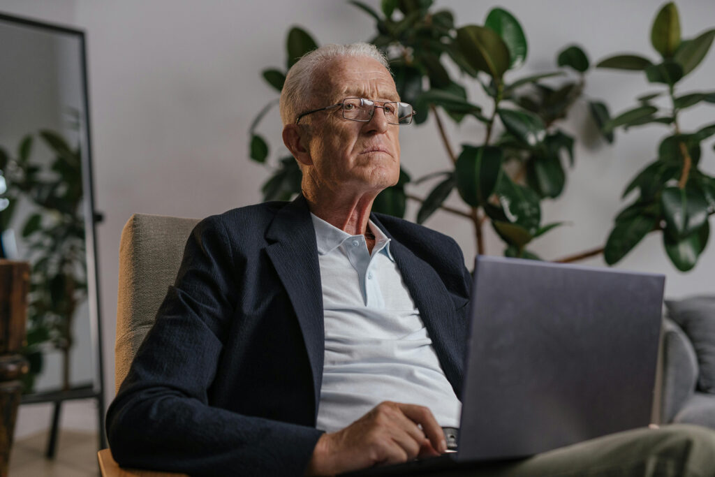 Ein älterer Mann mit Brille und Anzug sitzt auf einem Stuhl und benutzt einen Laptop. Im Hintergrund sind große grüne Pflanzen und ein Ganzkörperspiegel zu sehen, was darauf hindeutet, dass er sich selbst in dieser ruhigen Umgebung mit den Prinzipien der zusammensetzbaren UX beschäftigt.