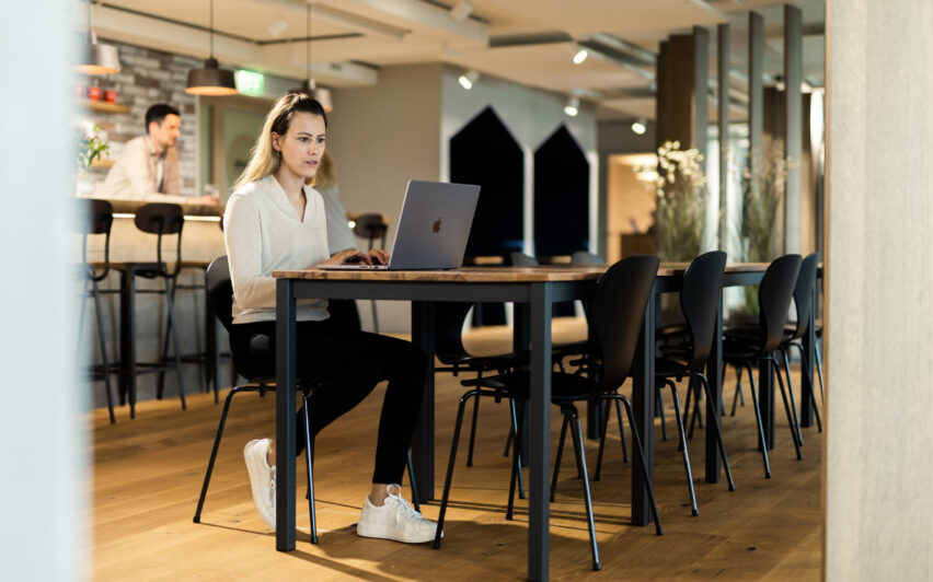 Eine Frau sitzt an einem langen Tisch und arbeitet an einem Laptop in einem modernen, offenen Büroraum. Im Hintergrund ist in der Nähe eines Barbereichs eine weitere Person zu sehen.