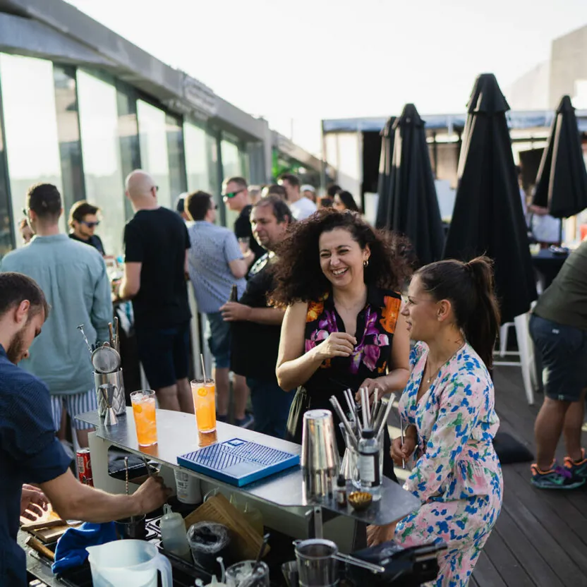 People are socializing and enjoying drinks at a rooftop bar during the daytime. The scene includes a bartender serving beverages and numerous patrons mingling around the bar.
