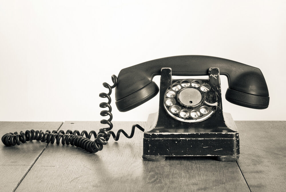 Ein altes schwarzes Telefon mit Wählscheibe steht auf einer Holzoberfläche vor einem schlichten Hintergrund.