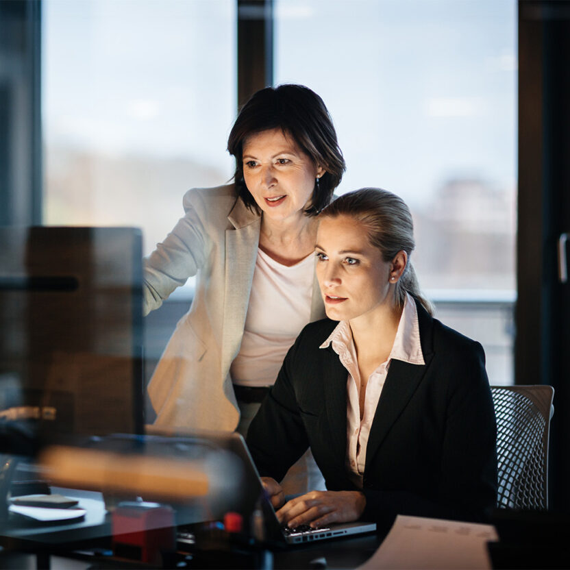 A young and mature businesswoman working late in front of a laptop and monitor in a modern office space. both faces are illuminated only by the computer monitor.