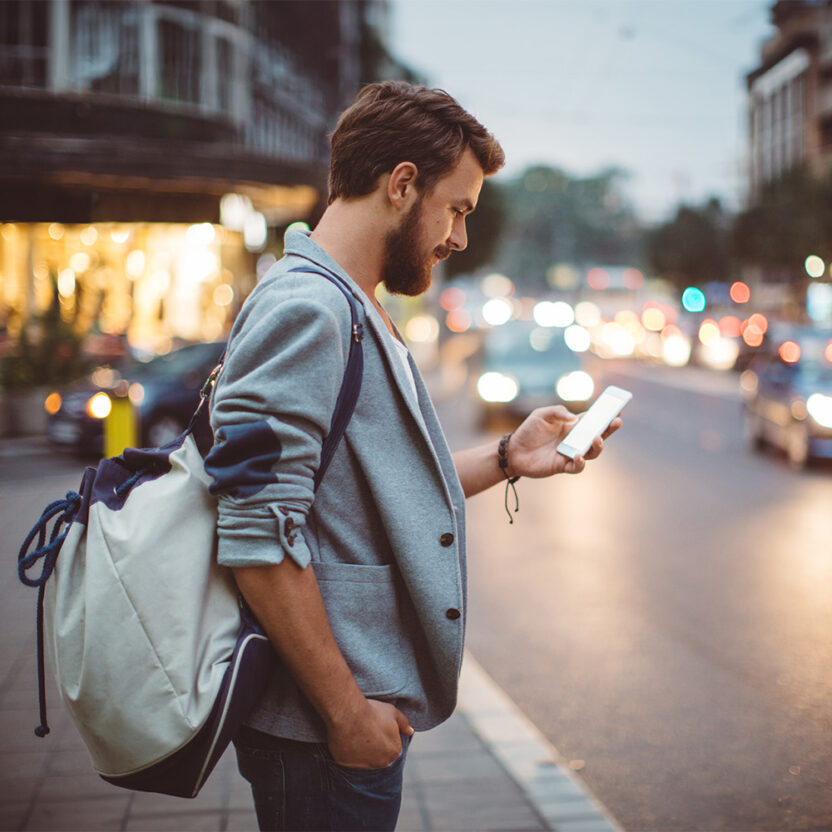 Young man on the street of big city , waiting taxi, chacking his smartphone, for news or new messages. Or looking for map instructions. Carry backpack on one shoulder. Dusk time. Casual dressed. Street lights in background.