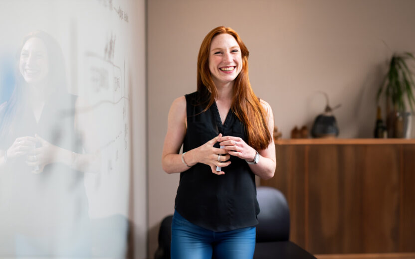 Woman in blue tank top standing beside white wall. Female software engineer in meeting by whiteboard.