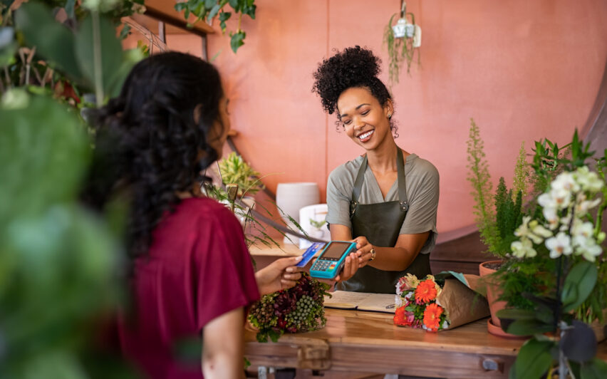 Smiling and friendly florist holding card reader machine at counter with customer paying with credit card.