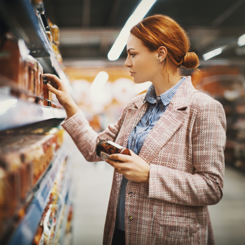 Young woman holding a jar of Sun-dried tomatoes in supermarket. She's searching for specific manufacturer.