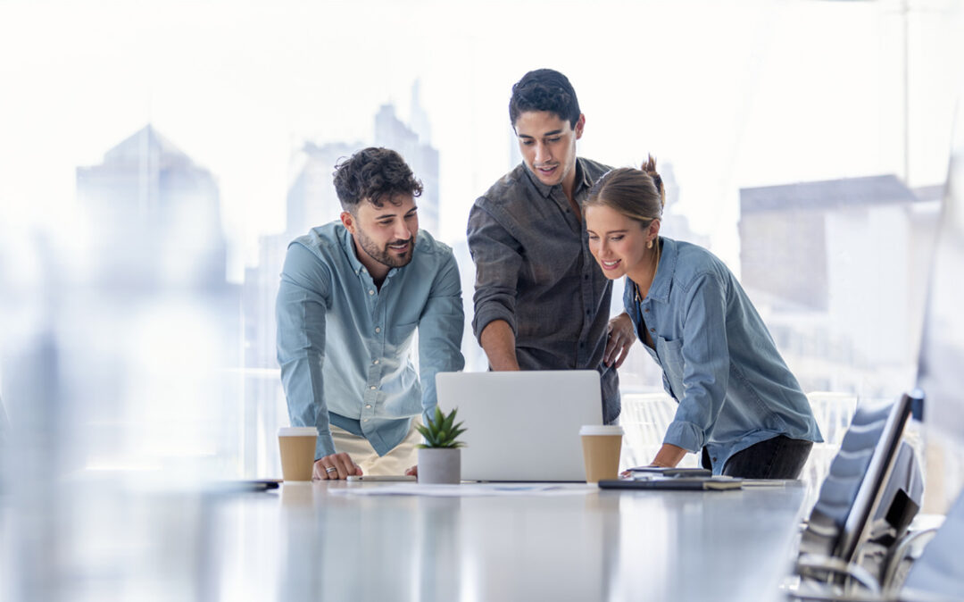 Business team working on a laptop computer. Three people are wearing casual clothing. They are standing in a board room. Multi ethnic group with Cauc and o men and women