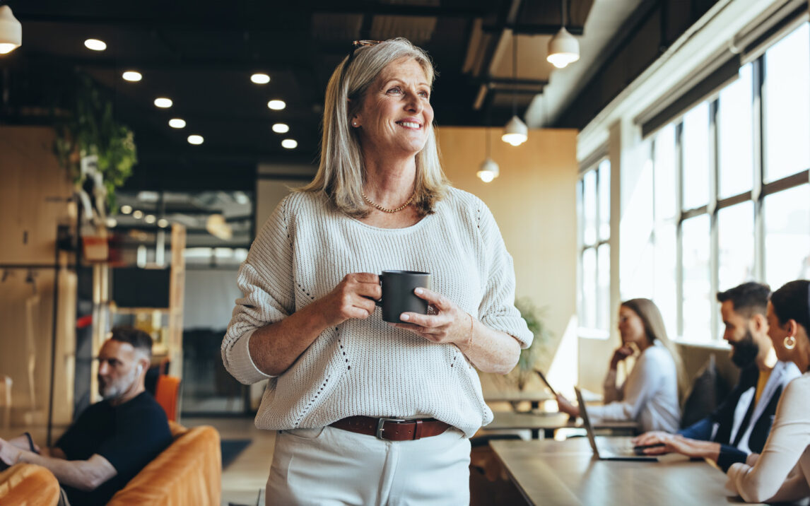 Thoughtful businesswoman standing in a co-working space. Mature businesswoman smiling cheerfully while holding a cup of coffee. Experienced entrepreneur contemplating new ideas during her coffee break.