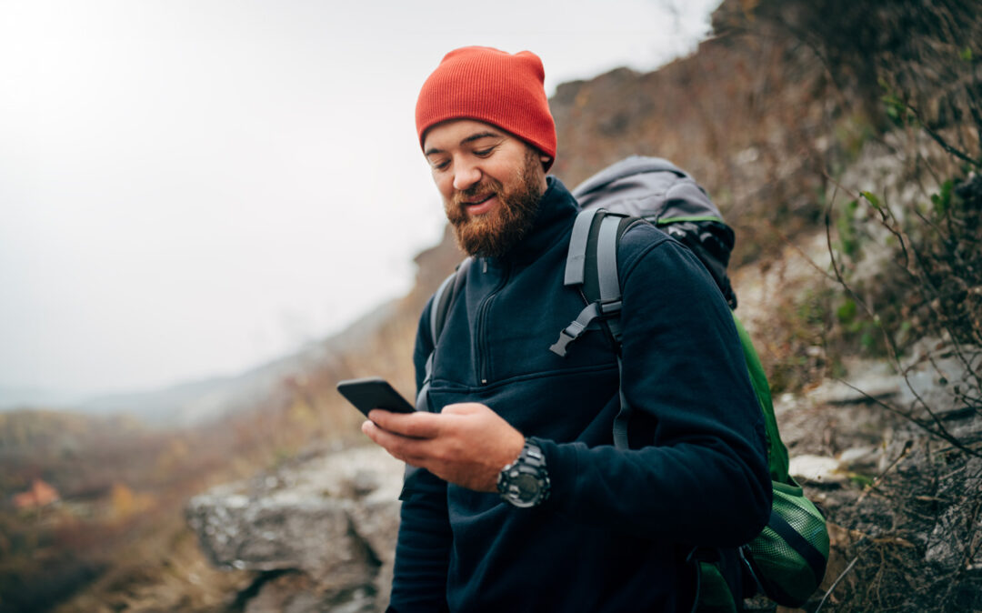 Young bearded man smiling and sending messages for his family from his cellphone, during hiking in mountains. Traveler bearded man in red hat using mobile phone application. Travel and lifestyle