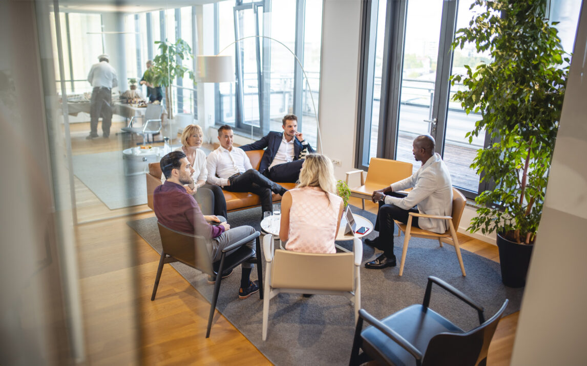 Elevated personal perspective photographed through window of diverse business colleagues relaxing and talking in modern office lobby.