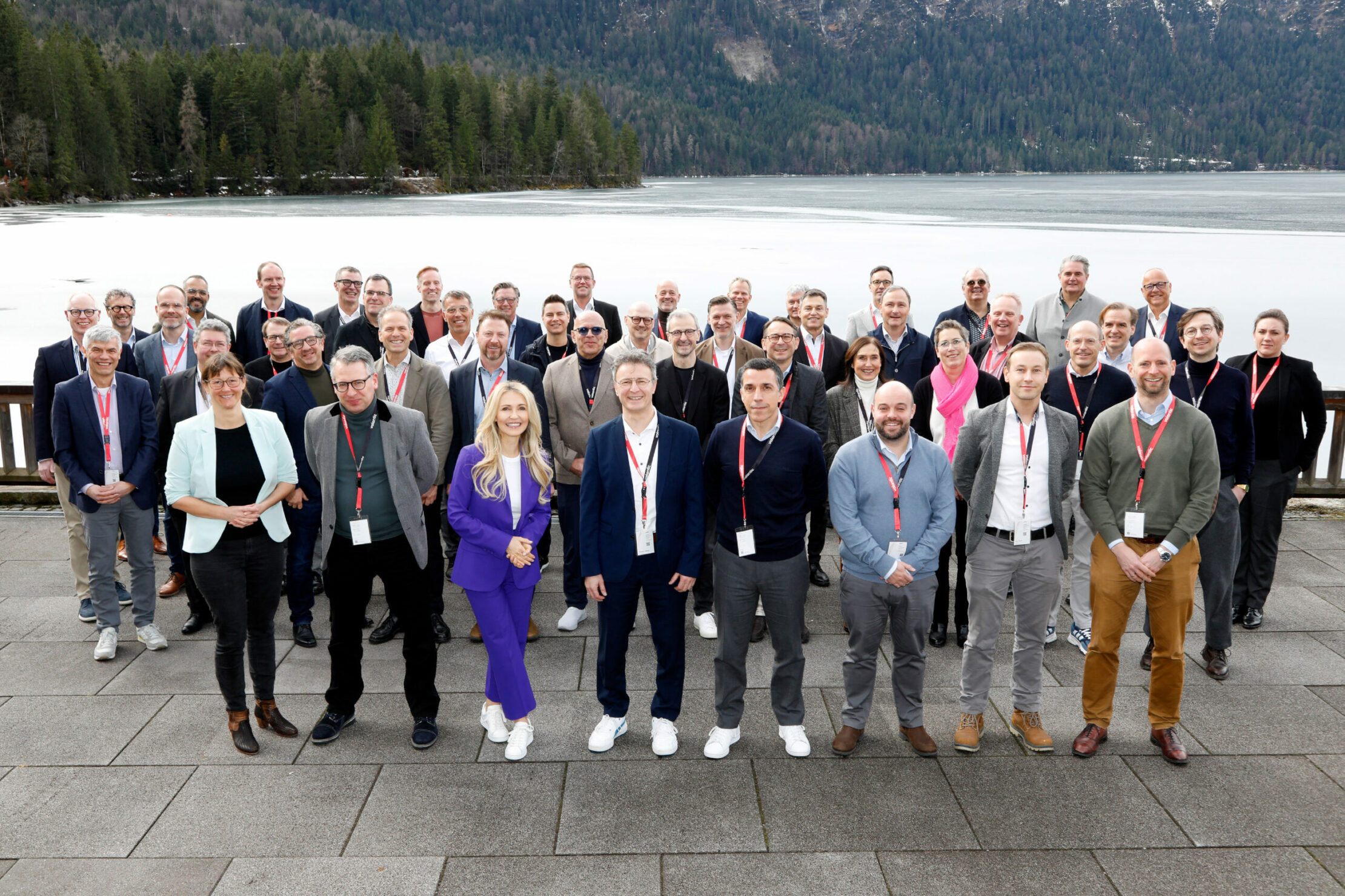 A group of people wearing conference badges stand on a patio with a scenic lake and forested mountains in the background.