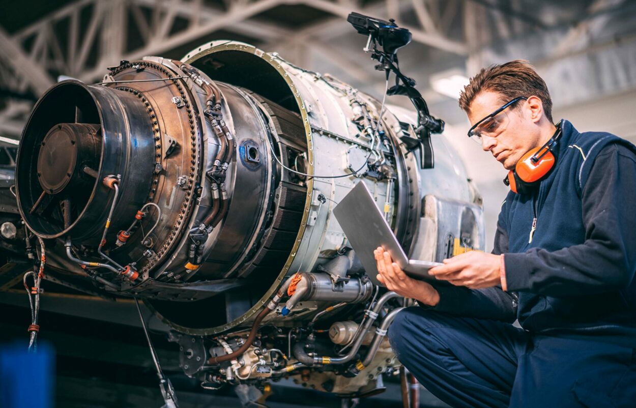 Aircraft engineer in a hangar using a laptop while repairing and maintaining an airplane jet engine