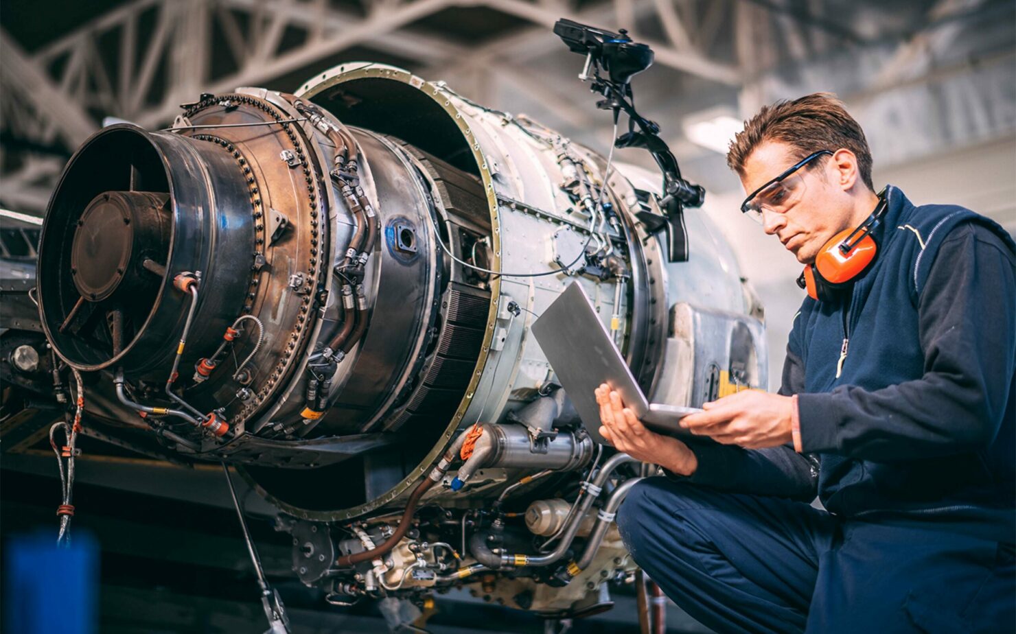 Aircraft engineer in a hangar using a laptop while repairing and maintaining an airplane jet engine