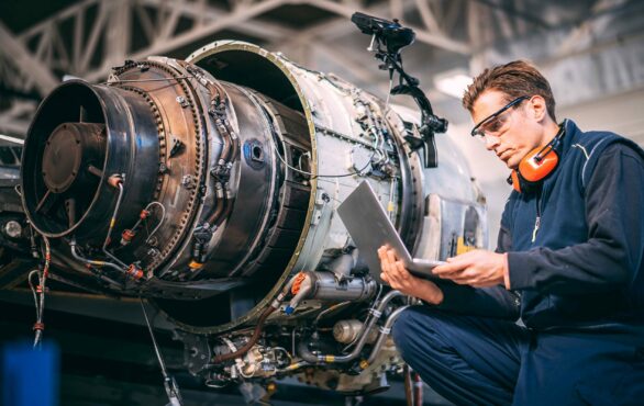 Aircraft engineer in a hangar using a laptop while repairing and maintaining an airplane jet engine