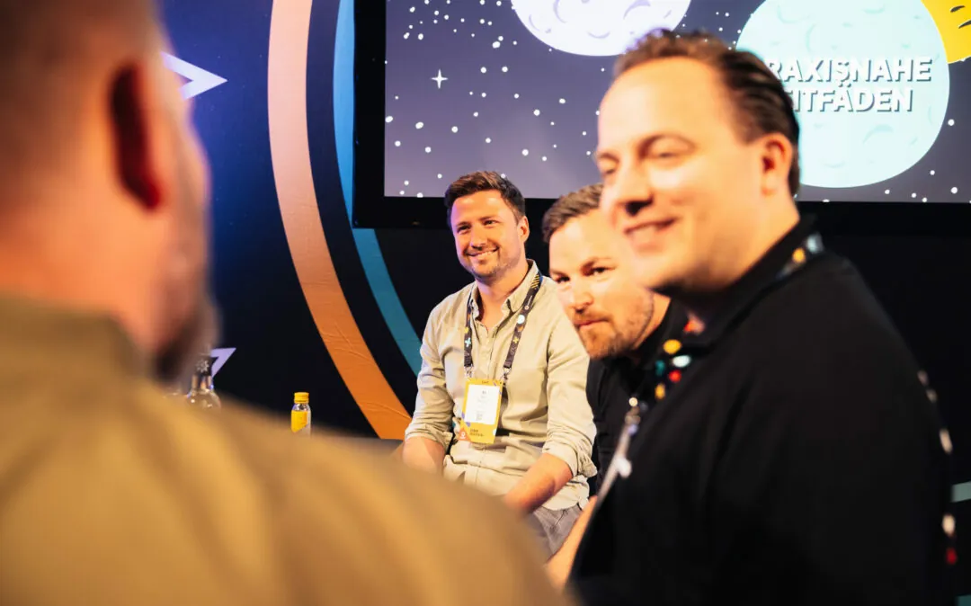 Eine Gruppe von vier Männern nimmt an einer Podcast-Sitzung vor einem Hintergrund mit einer Mondillustration und dem Wort „PODCAST“ teil. Drei Männer sind scharf zu sehen, während einer im Vordergrund unscharf ist.