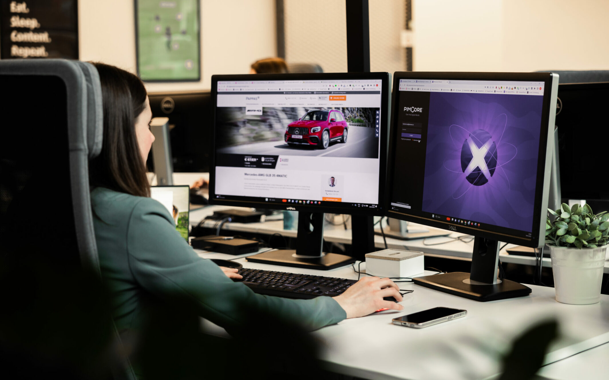 Eine Person sitzt an einem Schreibtisch und arbeitet an zwei Monitoren. Auf einem Bildschirm ist eine Autowerbung zu sehen, auf dem anderen ein violettes Design. Auf dem Schreibtisch liegen außerdem ein Smartphone und eine kleine Pflanze.