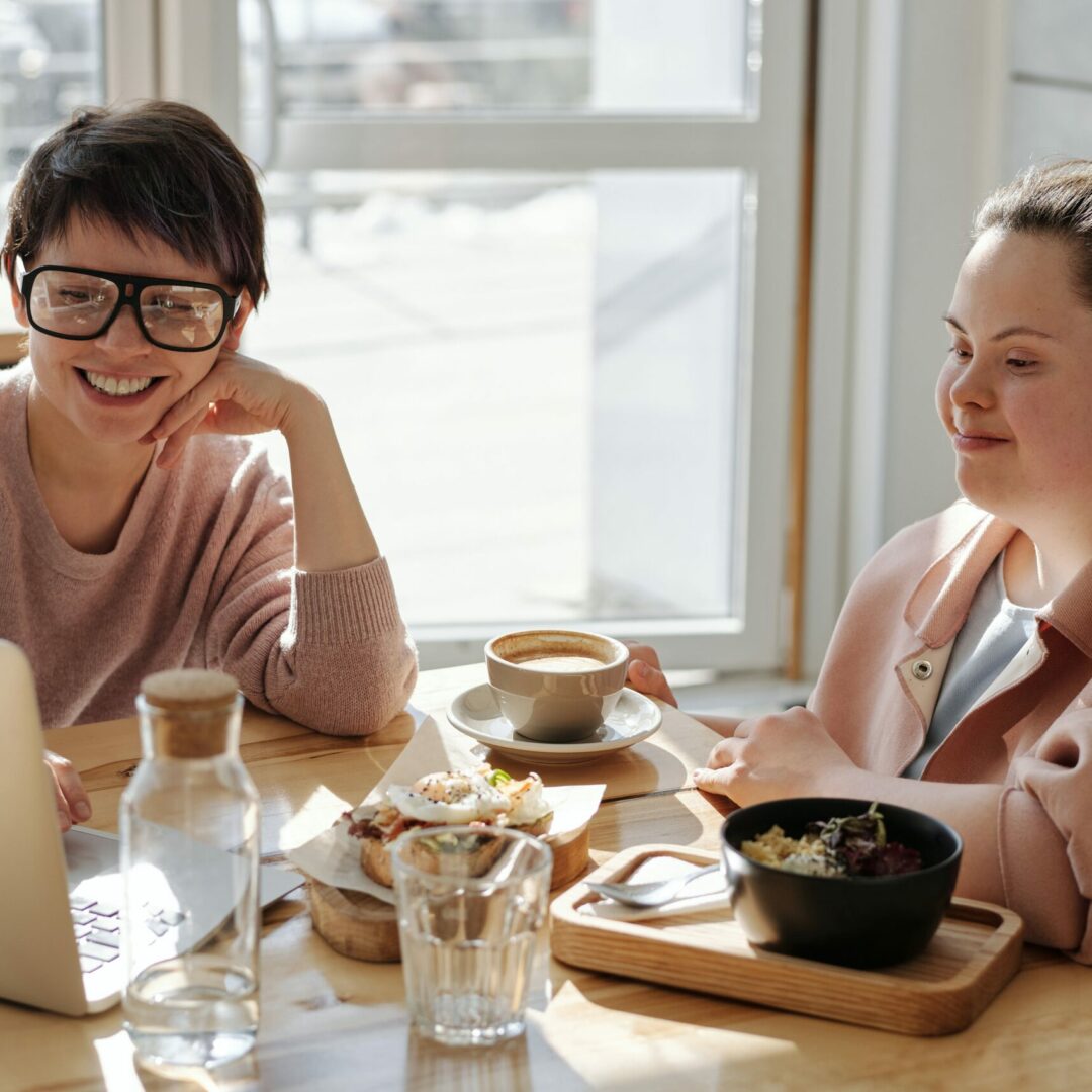 An einem Tisch mit Kaffee und Snacks sitzen zwei weibliche Personen, eine davon hat Down-Syndrom. Sie schauen lächelnd auf einen aufgeklappten Laptop.