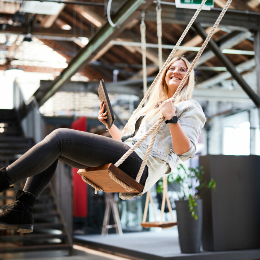 A woman holding a tablet swings on a wooden swing indoors, with industrial decor and a staircase in the background.