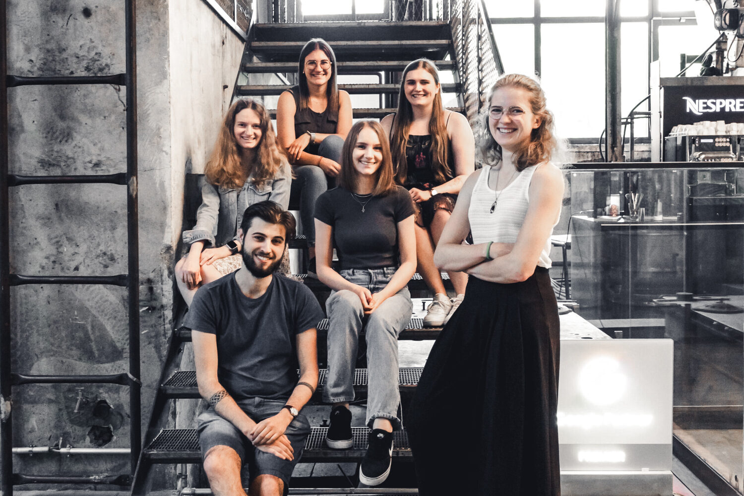 Vier junge Frauen und ein junger Mann sitzen auf einer Stiege und lächeln in die Kamera. Eine weitere junge Drau steht schräg links von der Stiege und hat ebenfalls ein Lächeln im Gesicht.