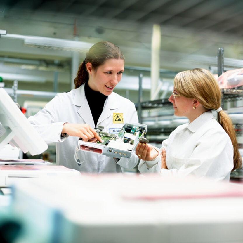 Two lab technicians in white coats discuss a Biotronik device in a laboratory setting. Biotronik valantic Supply Chain Excellence Day APS Software wayRTS