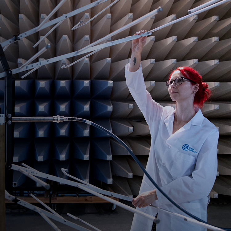 A person with red hair and glasses, wearing a white lab coat, adjusts equipment in an anechoic chamber with soundproofing foam panels.