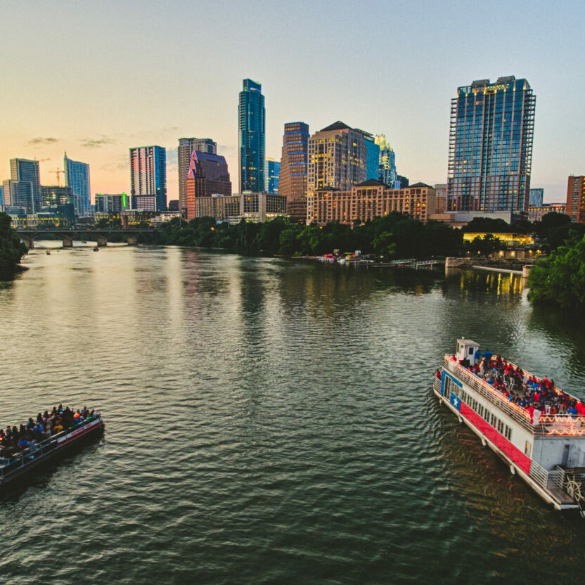 Two boats are cruising on a river at sunset with a city skyline featuring high-rise buildings in the background. Trees line the riverbanks. One boat is larger and brightly lit, while the other is smaller and darker.