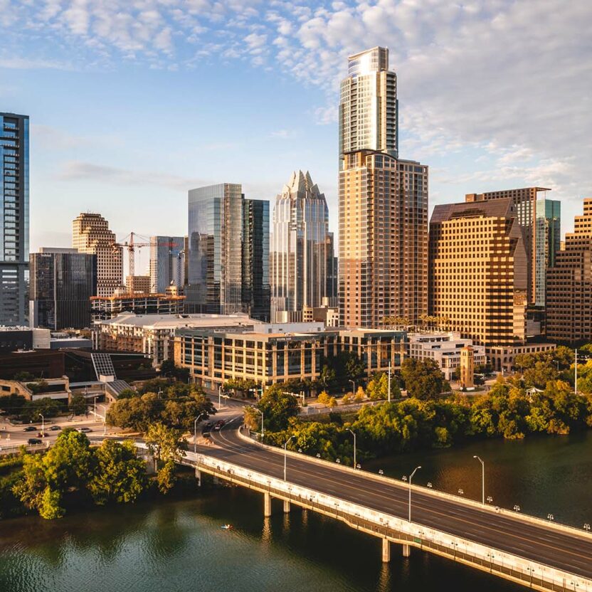 Skyline of downtown Austin, Texas at sunset with various high-rise buildings, a bridge crossing the river, and green areas along the waterfront.