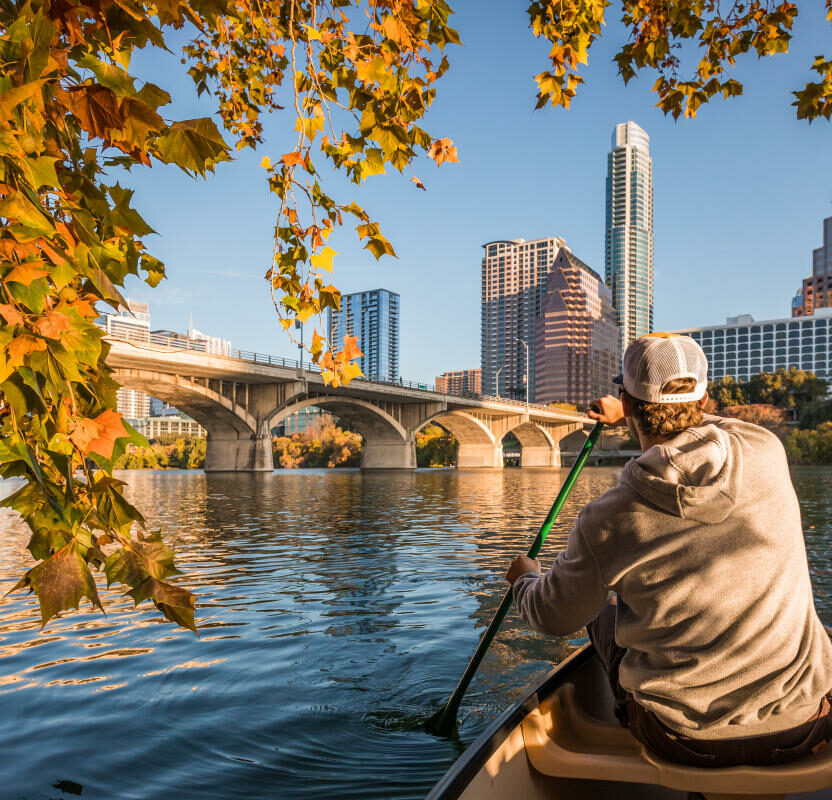 A person in a canoe paddles on a calm river, with a bridge and city skyline in the background, framed by autumn leaves.