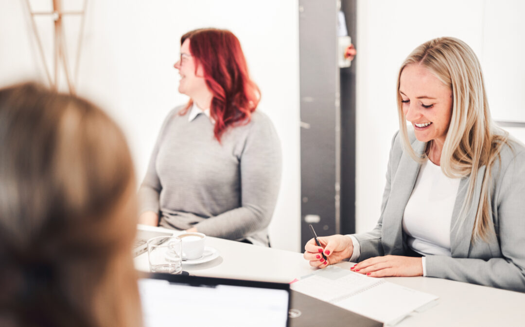 Drei Personen sitzen an einem Tisch in einem hellen Büro und sind in eine Besprechung vertieft. Eine Person benutzt einen Laptop, eine andere lächelt und schreibt und eine dritte blickt zur Seite.