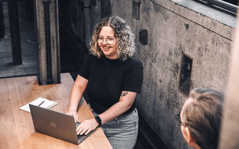 Eine Person mit lockigem Haar und Brille arbeitet an einem Laptop an einem Holztisch. Daneben liegt ein aufgeschlagenes Notizbuch, im Vordergrund ist eine weitere Person teilweise zu sehen.