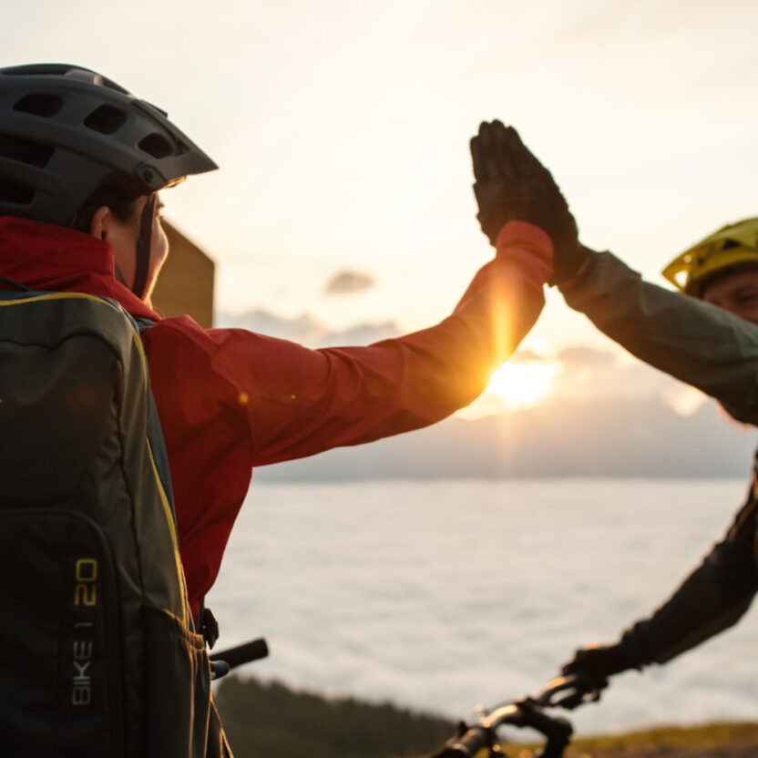 Zwei Mountain-Bike-Fahrer, die sich gerade High-Five geben - im Hintergrund sieht man den Sonnenuntergang.