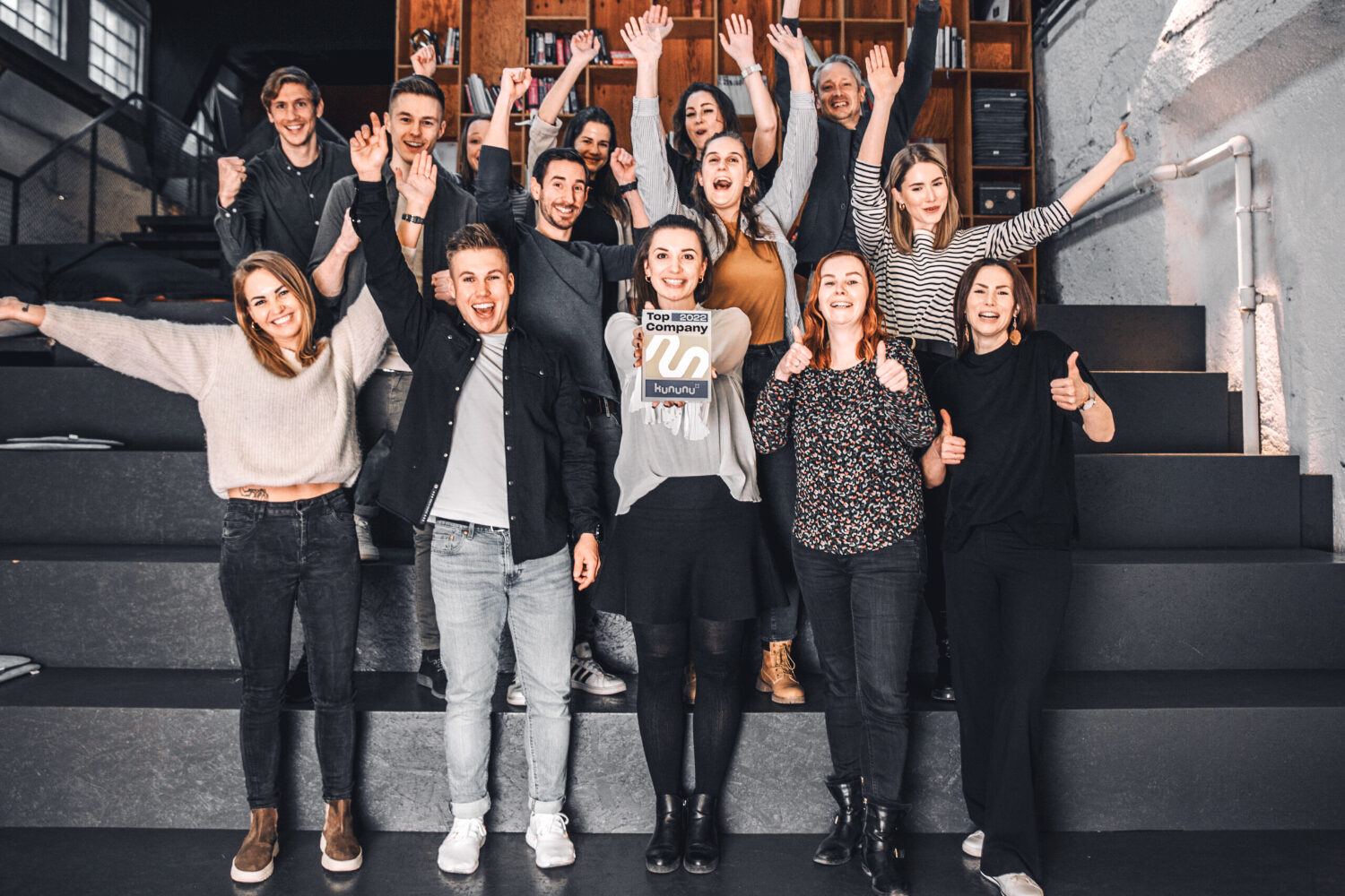 Foto von einer Gruppe mit 15 Personen, die auf einer Treppe stehen und jubeln. Eine Mitarbeiterin hält die Kununu Top Company Auszeichnung nach vorne.