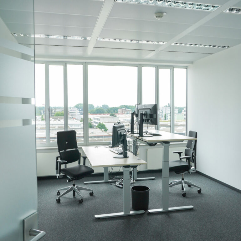 An office with two height-adjustable workstations, large windows and a gray carpet floor