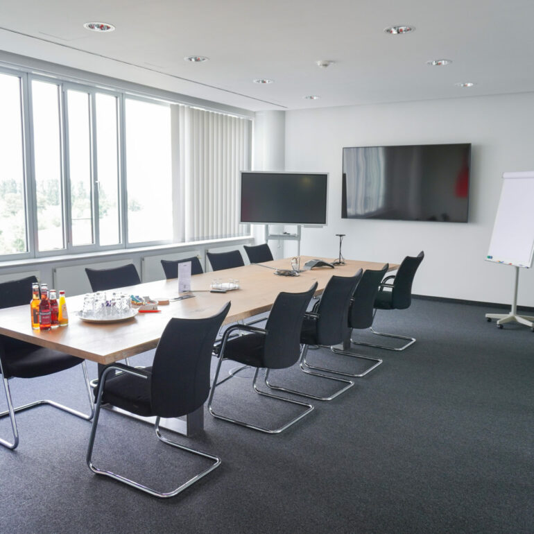 Bright meeting room with long conference table, gray carpeted floor, white walls and a large screen on the wall