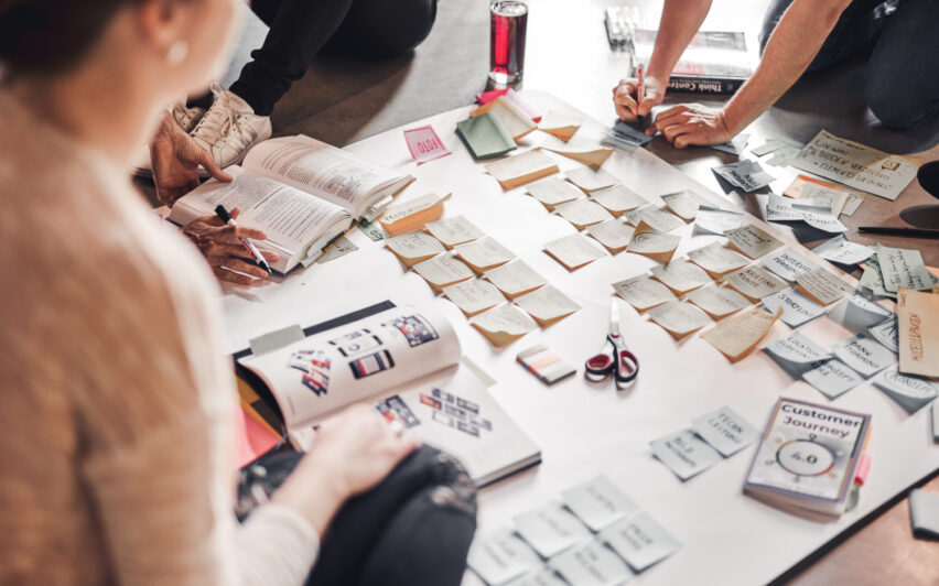 Mehrere Personen sitzen auf einem Boden um ein Plakat, auf das sie mit der Hand beschriftet Post-Its aufkleben und eine Customer Journey für einen Kunden von valantic Austria erarbeiten.