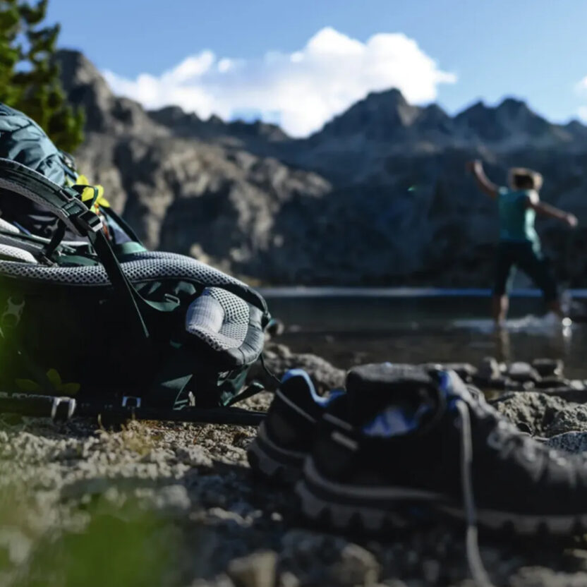 Foto von einem Deuter-Rucksack der auf einem Felse liegt. Im Hintergrund sieht man verschwommen Berge und einen Bergsee, sowie zwei Personen.