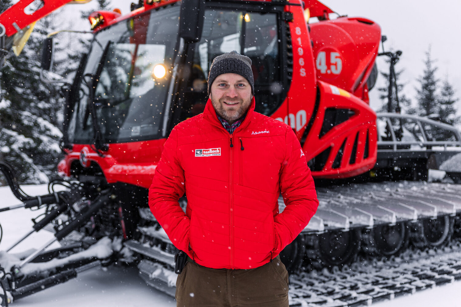 Der Pistenraupenfahrer Bartlmä trägt eine rote Skijacke und eine schwarze Mütze und steht vor einer roten Pistenraupe. Im Hintergrund sieht man einen beschneiten Wald.