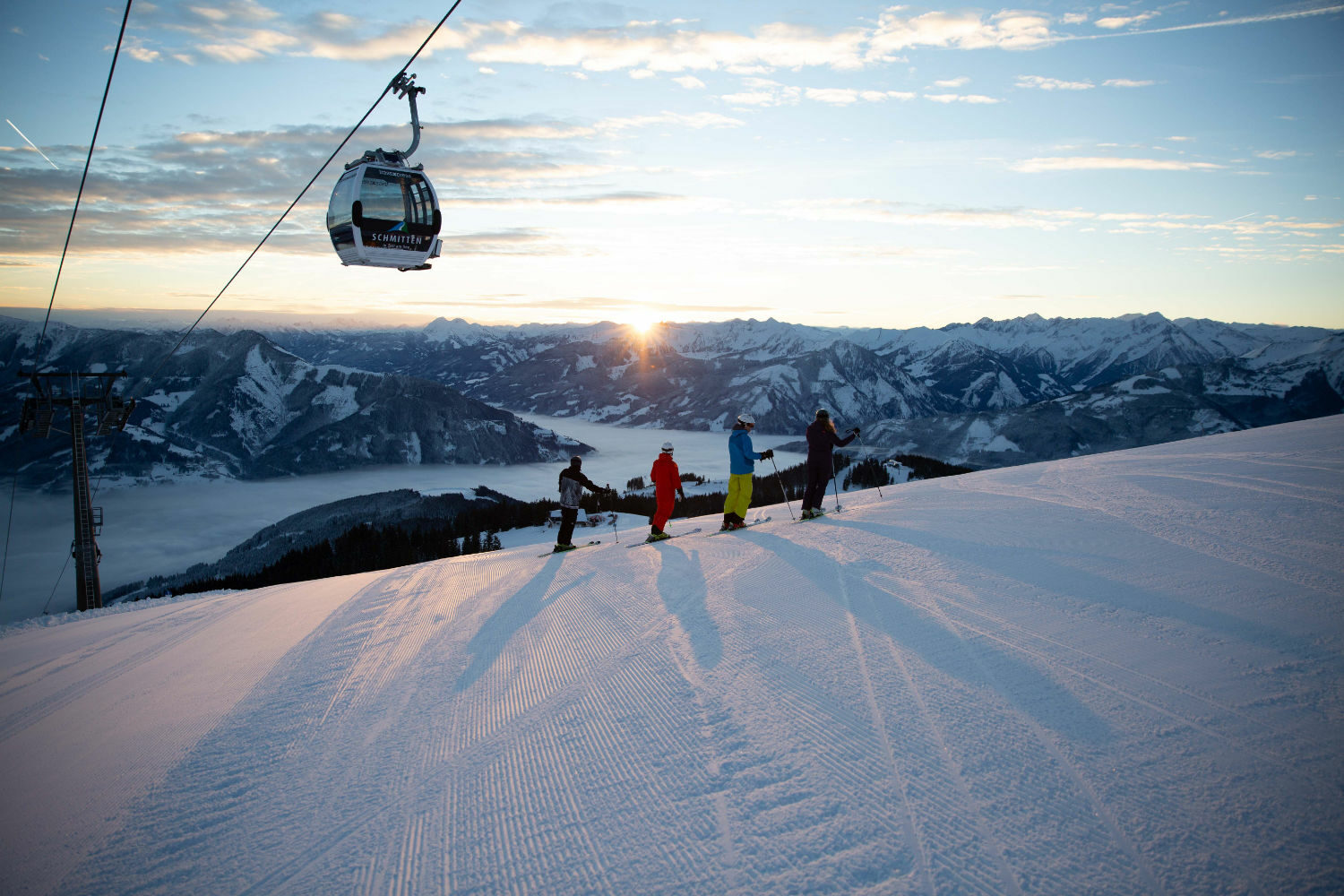 Vier Personen beim Schneeschuhwandern bei Sonnenuntergang. Über ihnen schwebt eine Gondel der Schmittenhöhenbahn.